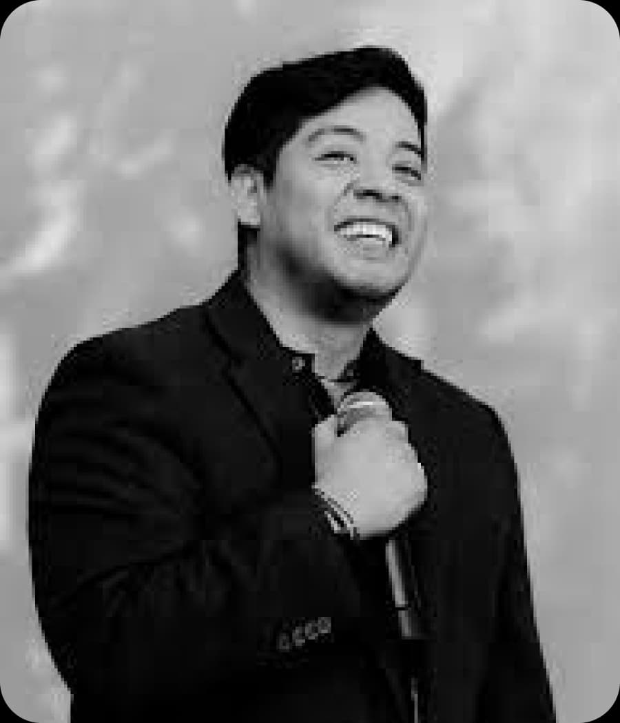 Man in a suit jacket laughing while holding a microphone.